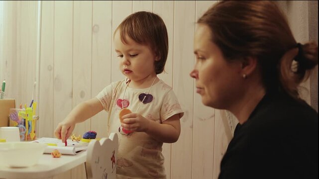 Little girl explores colorful clay at a play table while her mother stays close and supports her creativity.