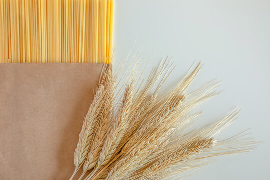 Raw dry spaghetti in kraft paper packaging placed beside golden wheat stalks on a clean white background. Concept of wheat processing, pasta manufacturing and farm to table production. 