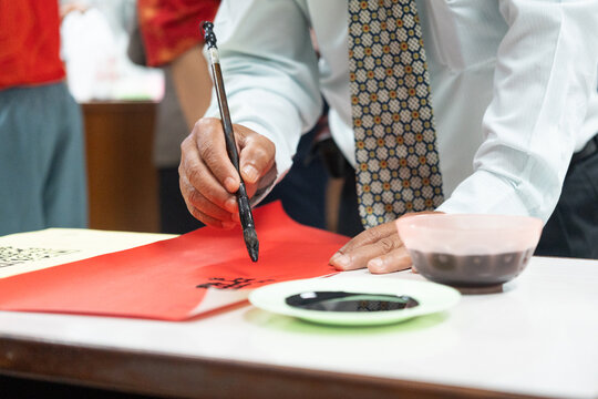 Focused student in blazer writing traditional Chinese calligraphy characters with ink brush on vibrant red paper, demonstrating authentic cultural heritage and tactile craftsmanship