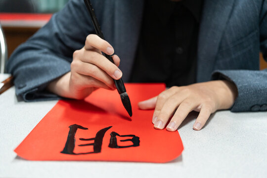 Focused student in blazer writing traditional Chinese calligraphy characters with ink brush on vibrant red paper, demonstrating authentic cultural heritage and tactile craftsmanship