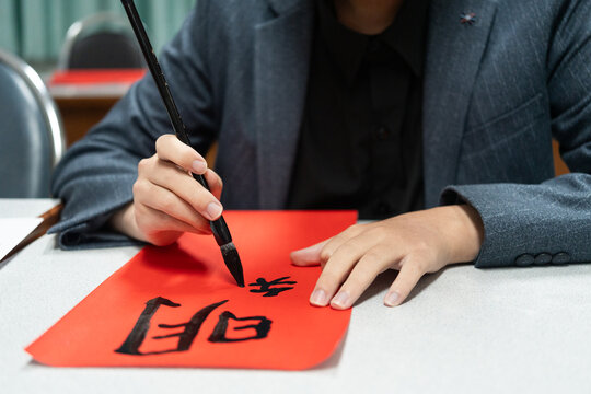 Focused student in blazer writing traditional Chinese calligraphy characters with ink brush on vibrant red paper, demonstrating authentic cultural heritage and tactile craftsmanship