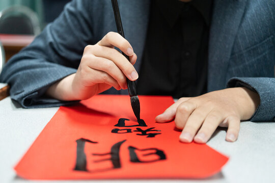 Focused student in blazer writing traditional Chinese calligraphy characters with ink brush on vibrant red paper, demonstrating authentic cultural heritage and tactile craftsmanship