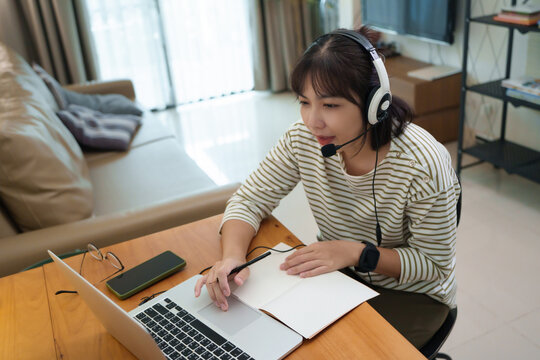 Young student attends an online class with a headset while taking notes on a laptop at home, highlighting remote learning, digital education, and active participation.
