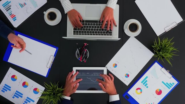 Business team works with financial documents and a tablet presentation at an office desk, taking notes while planning a project in a flat lay workspace.