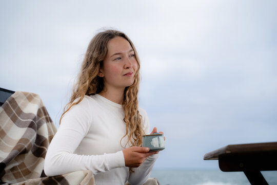 Woman enjoying a hot drink by the ocean while sitting outdoors in cool weather. Calm coastal atmosphere, mindfulness and relaxation. Side view portrait.
