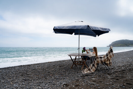 Cozy seaside setup with a woman enjoying nature under an umbrella on a rocky beach. Mindful relaxation, leisure and coastal escape.