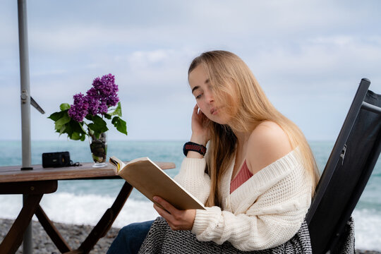 Young blonde woman enjoying a printed book while sitting at a table by the sea. Peaceful coastal recreation, slow living and mindful outdoor lifestyle.