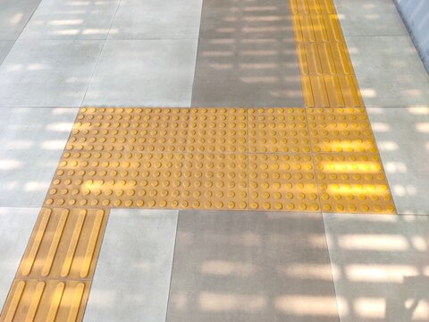 Yellow tactile paving tiles with raised dots and lines form a path on a grey tiled floor under dappled shadows.