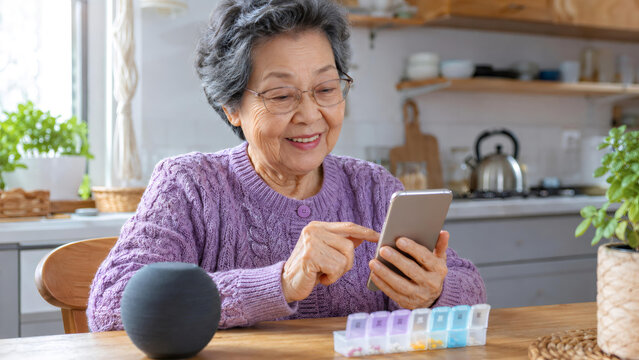 Asian woman using smartphone to manage daily medication schedule at home. Senior lady with pill organizer and smart assistant device. Health tracking and technology for elderly care.