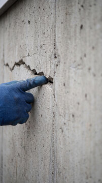 Inspector hand in glove pointing at deep crack in grey concrete wall. Structural damage assessment in building construction. Maintenance engineer checking wall integrity and safety hazard.