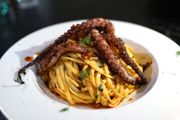 A plated Italian pasta dish featuring grilled octopus tentacles over linguine in chili garlic oil, garnished with fresh herbs. © ltyuan
