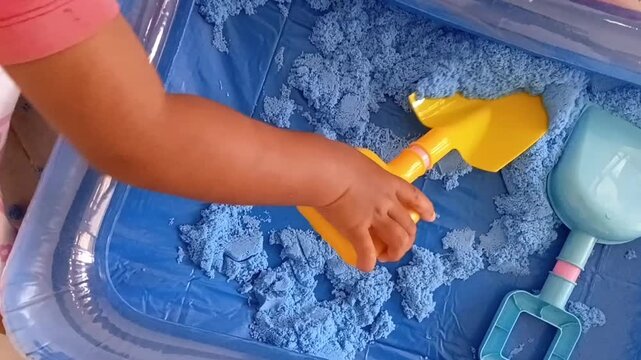 Close-up video of a child's hand playing with blue kinetic sand in a plastic tray. The toddler uses a yellow shovel and pink crab mold for sensory play.