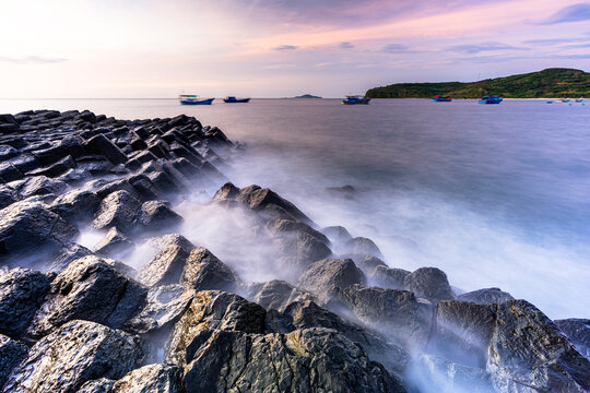Waves crash against volcanic rocks at Da Dia Reef in Phu Yen Vietnam during sunset with fishing boats in the background