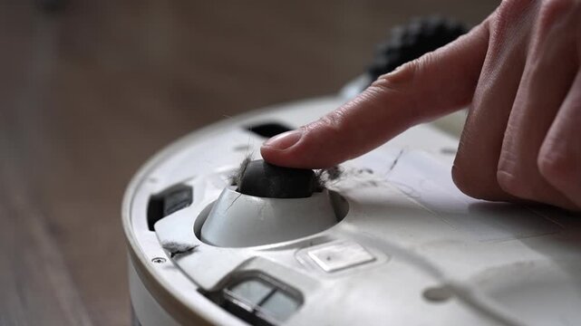 Close-up of person cleaning hair, dust, debris from dirty caster wheel of robotic vacuum cleaner. Hands remove, clean, reinstall part. Home appliance maintenance and repair concept, slow motion.