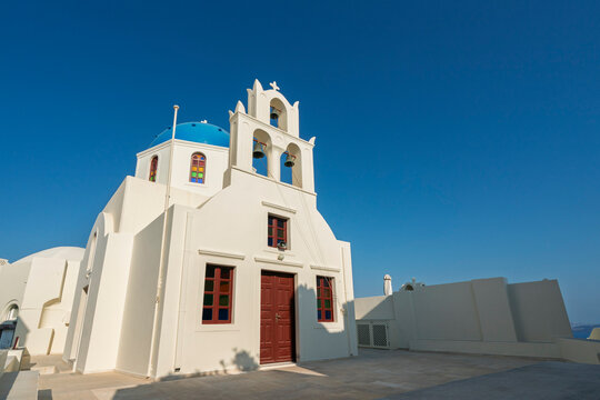 Church with blue dome stands on sunny day in Oia, Santorini Island, Cyclades archipelago, Greece while visitors stroll nearby