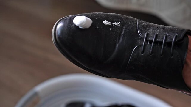 Male hand applying special white foam cleanser from bottle onto classic black leather shoe and using sponge to scrub and clean entire surface. Concept of professional footwear maintenance.