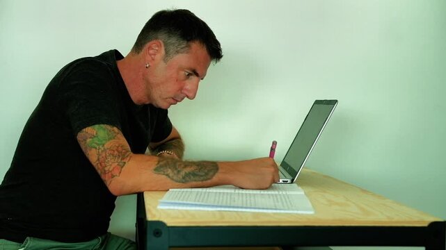 A man studies in front of a computer and a notebook while sitting at a desk. The man is forty-two years old and wears a dark T-shirt. Video shows adult student's efforts
