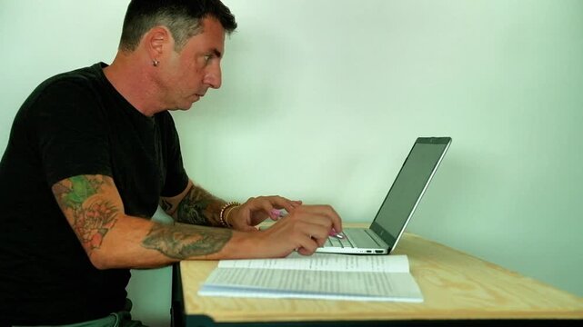 A man studies in front of a computer and a notebook while sitting at a desk. The man is forty-two years old and wears a dark T-shirt. Video shows adult student's efforts
