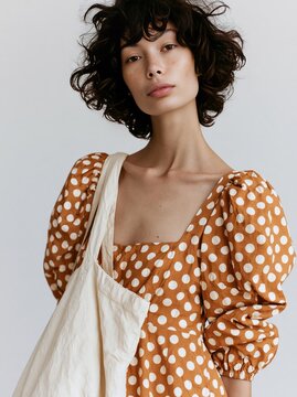 Woman wearing a brown polka dot midi dress with puff sleeves, holding a beige tote bag, against a neutral background, showcasing modern fashion and style