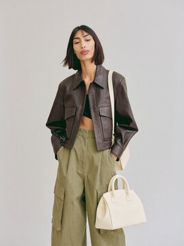 Woman wearing a dark brown leather jacket and olive green cargo pants stands confidently with a small cream handbag against a neutral background