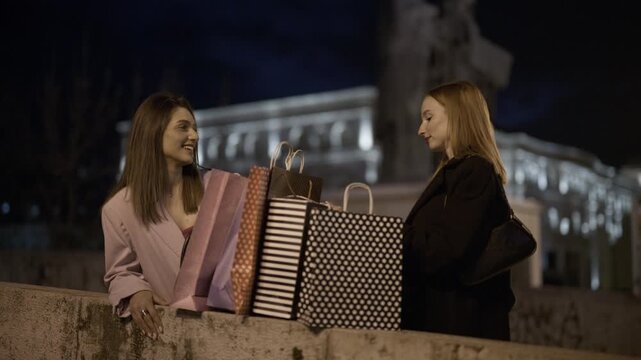 Two happy girls stand with shopping bags in a nighttime city. They are smiling and talking, enjoying their recent purchases.