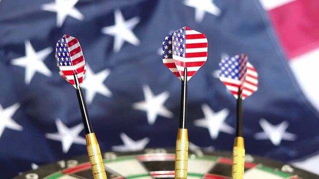 Three metal darts featuring American flag patterns stand firmly in the center of a dartboard with a large United States flag hanging in the background during a patriotic holiday celebration event