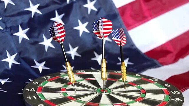 Three darts decorated with the American flag patterns hit the target on a dartboard while the stars and stripes flag waves behind them indoors