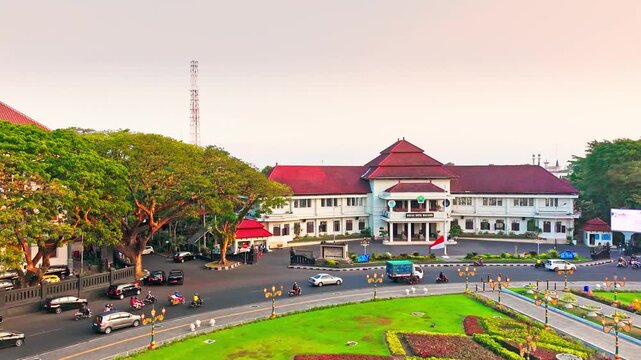 Golden hour bathes historic building and central monument. Circular pond reflects sky and structure. Lush greenery surrounds the scene. Distant trees frame the peaceful vista