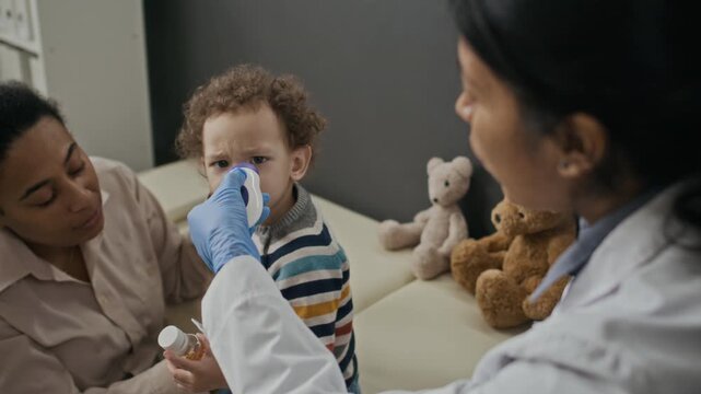 Hispanic female pediatrician measuring temperature of toddler boy with infrared thermometer during medical checkup with Biracial mother. Perfect for pediatric healthcare marketing campaigns