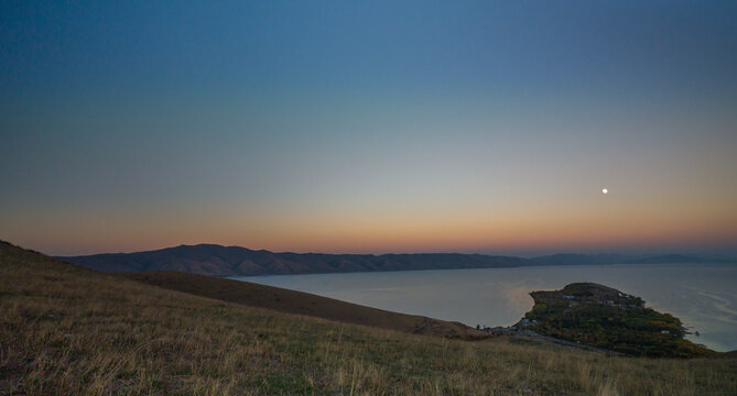 A peaceful evening view of Lake Sevan, with the full moon illuminating the still water and mountain ridges under the fading gradient of dusk.