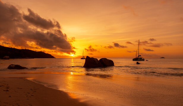 A picturesque beach scene in warm orange red hues featuring boats, rocks and calm water illuminated by sunset sky. Coastal landscape of Mahe island, Seychelles