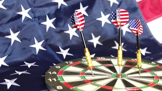 Three darts with patterns of the United States flag stand firmly in the center bullseye of a dartboard with a large American flag draped in the background during a focused afternoon hobby session
