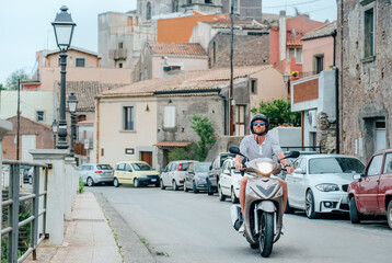 Happy tourist wearing helmet riding modern scooter through narrow historic street of Forza d'Agro village in Sicily exploring charming traditional Italian architecture sunny summer vacation travel day © Soloviova Liudmyla