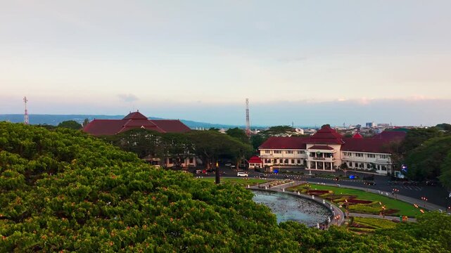 Golden hour bathes historic building and central monument. Circular pond reflects sky and structure. Lush greenery surrounds the scene. Distant trees frame the peaceful vista