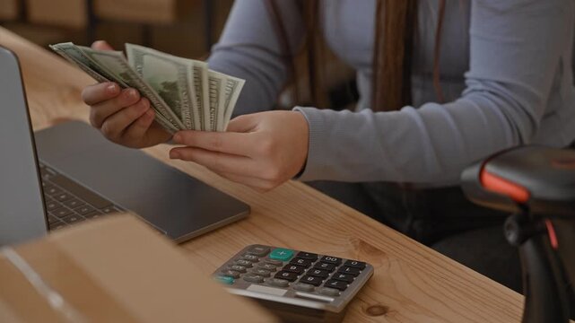 Woman counting cash with hands at a shipping desk in an office, using laptop, calculator and barcode scanner; small business earnings focus.