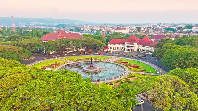 Golden hour bathes historic building and central monument. Circular pond reflects sky and structure. Lush greenery surrounds the scene. Distant trees frame the peaceful vista