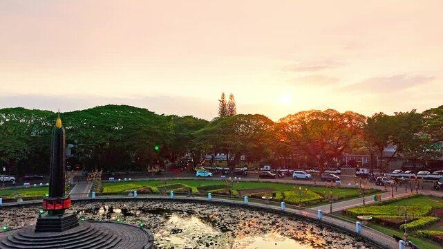 Golden hour bathes historic building and central monument. Circular pond reflects sky and structure. Lush greenery surrounds the scene. Distant trees frame the peaceful vista