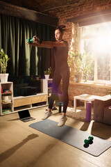 Obraz premium Athletic woman jumping with dumbbell during high intensity home workout. Concept of cardio strength training, calorie burning, online fitness and active lifestyle.