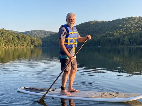 senior man paddleboarding lake fitness outdoors retirement balance adventure wellness A paddler stands on water near green hills showing freedom confidence and healthy aging for recreation campaigns