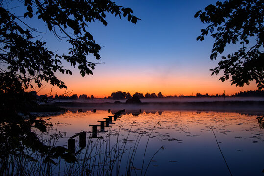 Misty lake at sunrise with old wooden pier posts, calm reflections, silhouetted trees, and a colorful dawn sky.