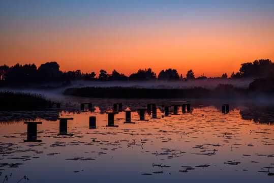 Misty lake at sunrise with old wooden pier posts, calm reflections, silhouetted trees, and a colorful dawn sky.