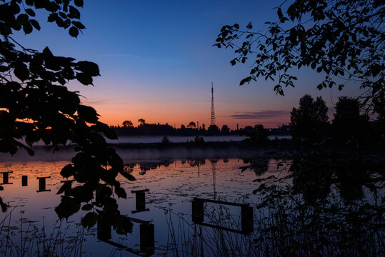 Misty sunrise over a calm lake with a tall radio tower, soft reflections, silhouetted trees, and colorful dawn sky.