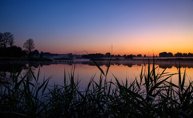 Obraz premium Misty lake at sunrise with old wooden pier posts, calm reflections, silhouetted trees, and a colorful dawn sky.
