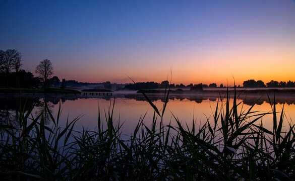 Misty lake at sunrise with old wooden pier posts, calm reflections, silhouetted trees, and a colorful dawn sky.