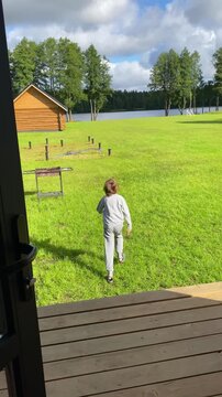 Vertical video of young boy running joyfully from wooden home across vibrant green lawn towards lake. Freedom, summer vacation, and childhood happiness.