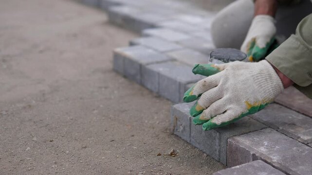 Gloved construction worker sets gray paving blocks with mallet beside compacted ground on outdoor jobsite, showing careful alignment, dusty texture, muted tones, and hands-on
