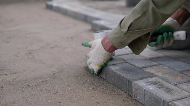 Gloved worker hand places dark gray paving blocks along straight roadside border on sandy ground, beside stacked pavers, rough curb pieces, muted light, and focused outdoor