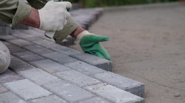 Protective gloved hands position gray paving blocks along curb edge on outdoor walkway, with rubber mallet resting across surface, stacked pavers, sandy base, blurred greenery