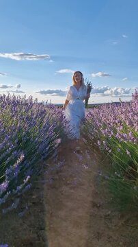 Beautiful young European woman with blonde hair wearing a white dress, holding a bouquet of lavender, walking and smiling in a scenic lavender field at sunset.