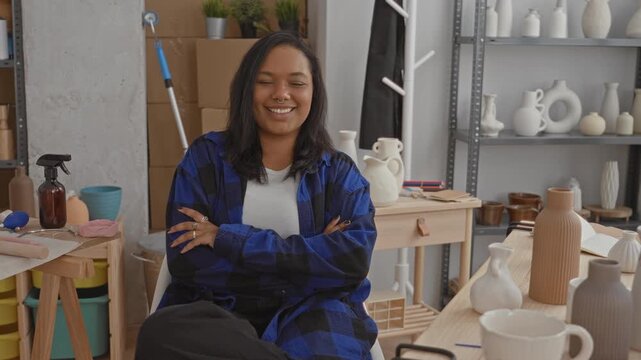 Young woman smiling with arms crossed among ceramic vases and pottery tools in a studio; creativity contentment.